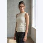 Woman wearing a beige sleeveless top and dark pants in a softly lit room.
